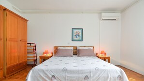 A bedroom with a wooden bed, two bedside tables with lamps, a wooden wardrobe, a wall-mounted air conditioner, and a framed painting on the wall.