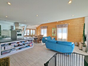 A modern living room with a blue sofa, a dining area with a table and chairs, a kitchen with a refrigerator and oven, and wooden walls.