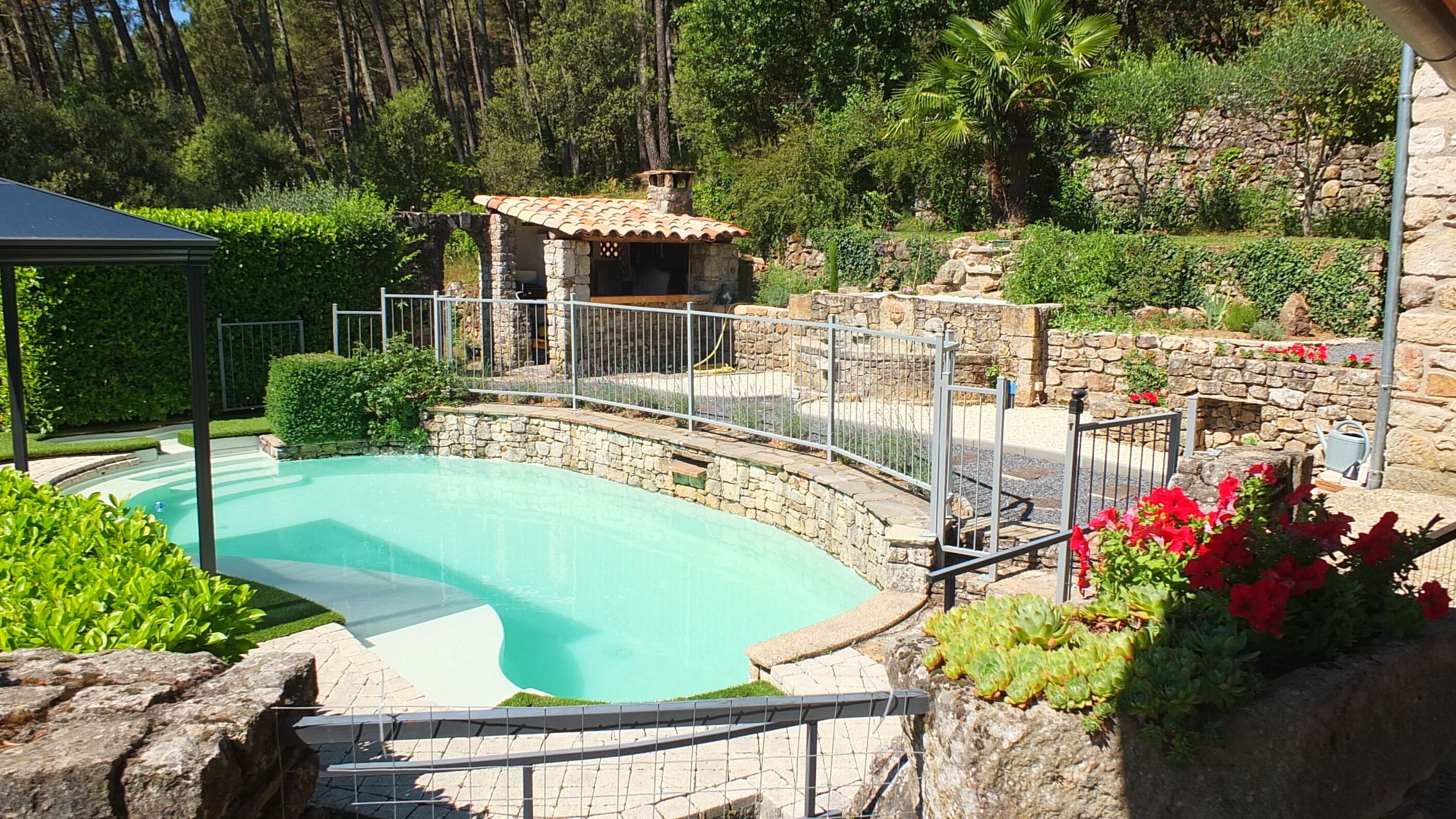 Une piscine dotée d’une plage en pierre, entourée d’un mur de pierre et d’une clôture en métal.