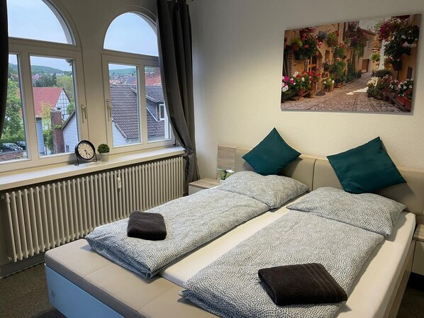 A bedroom with two beds, a window with a view of houses, a radiator, and a clock.