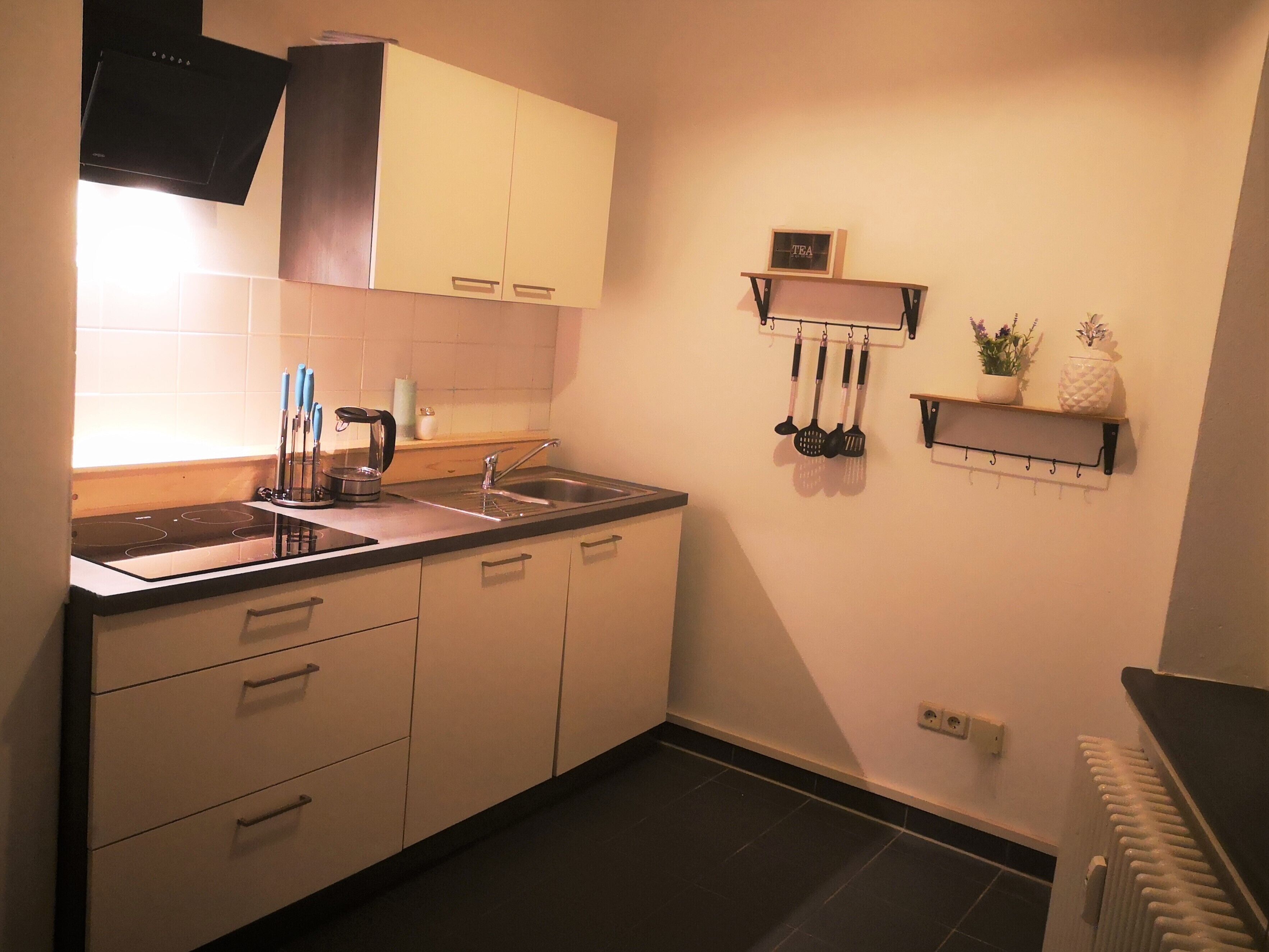 A compact kitchen with white cabinets, a black countertop, a stainless steel sink, and a wall-mounted rack holding kitchen utensils.