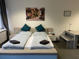 A double bed with two pillows, a bedside table with a lamp, a bench, and a framed picture on the wall.