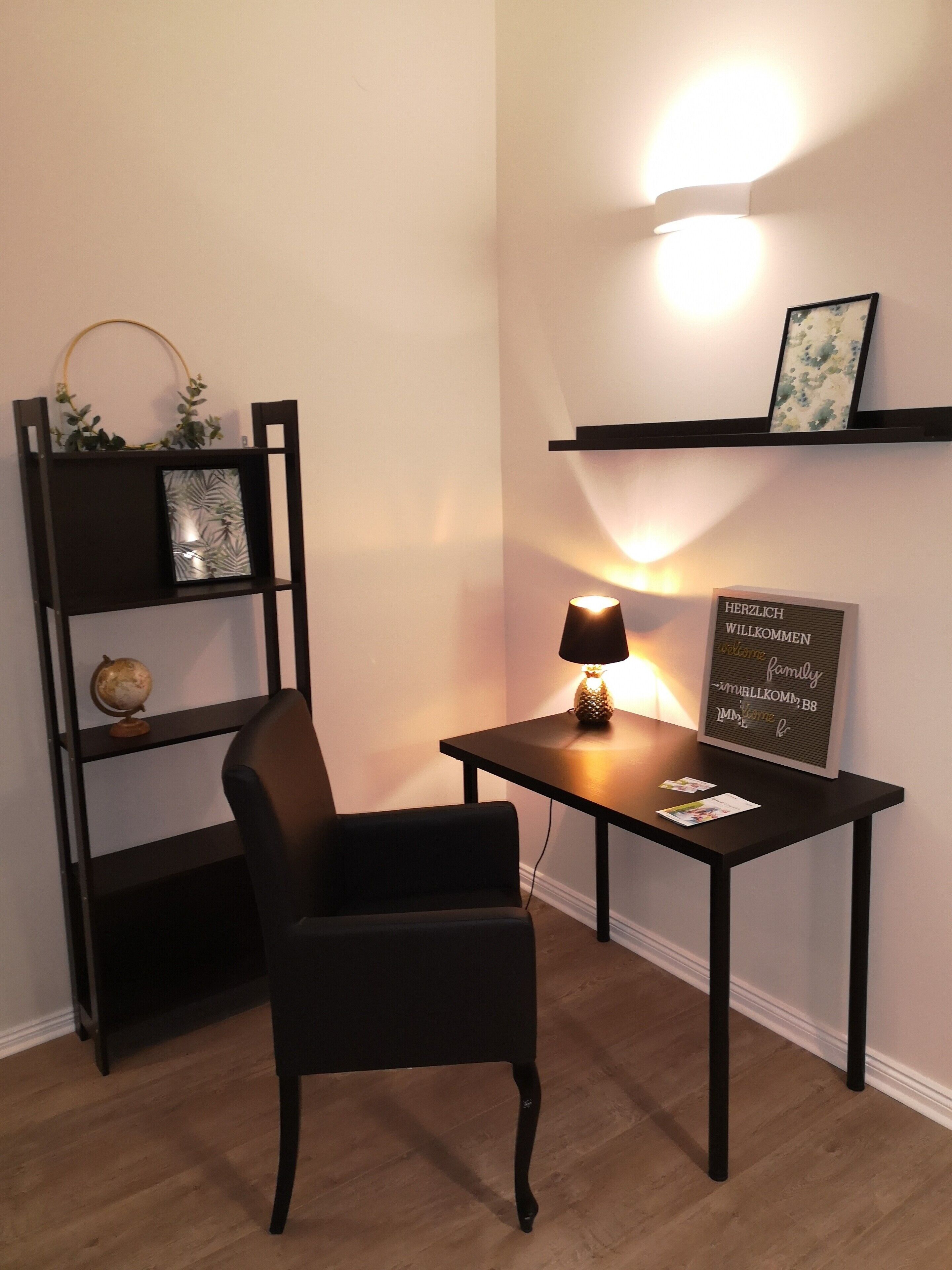 A small office space with a desk, chair, bookshelf, and a wall-mounted shelf with a framed welcome sign.