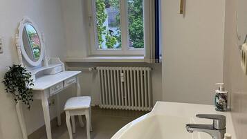 Een badkamer met een wit wastafelmeubel, een grote wastafel, een spiegel, een raam met uitzicht op groen en een radiator.