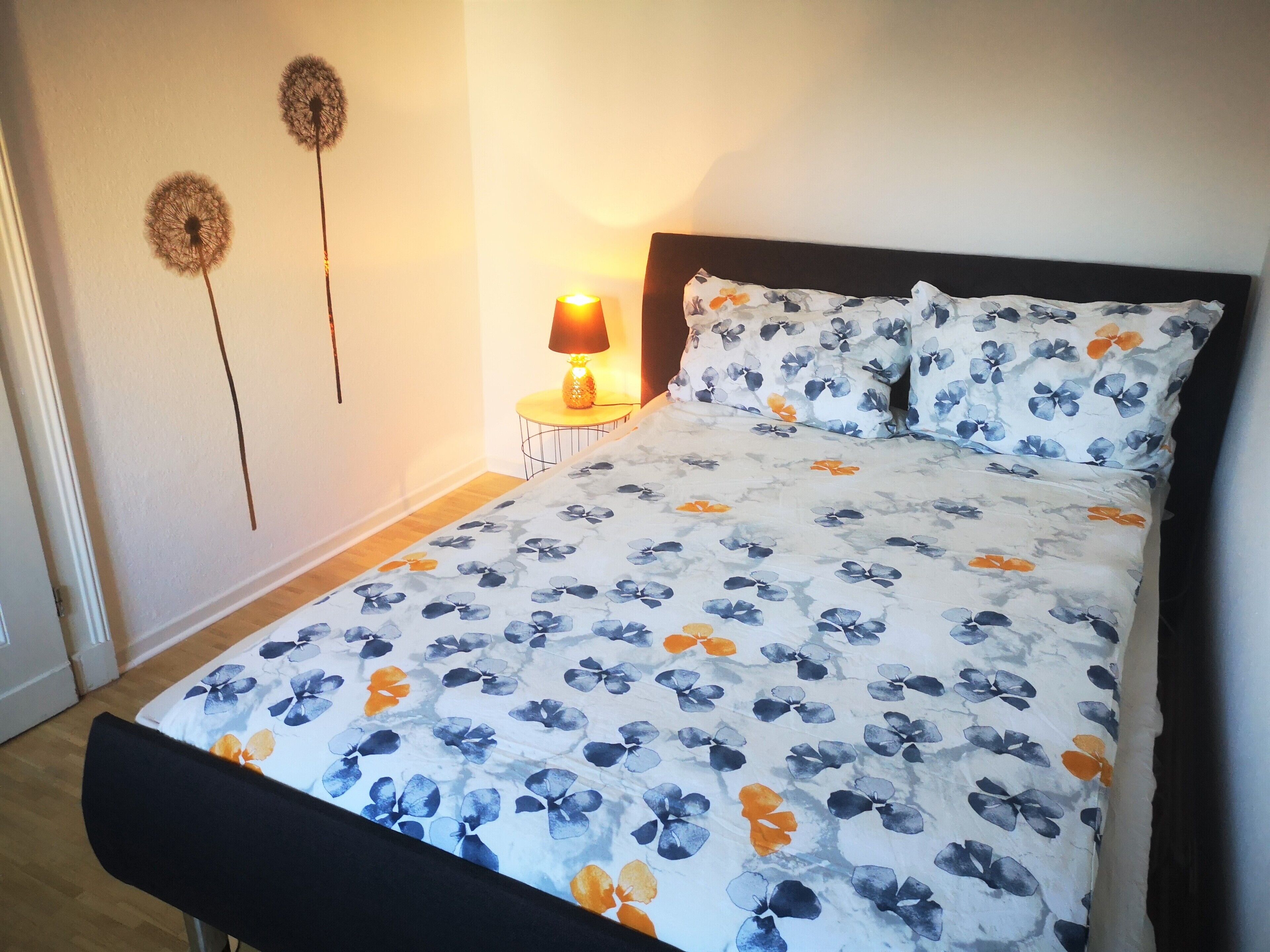 Un lit bien fait, recouvert d’une couvre-lit à motifs floraux, avec une lampe de chevet et deux éléments décoratifs accrochés au mur.