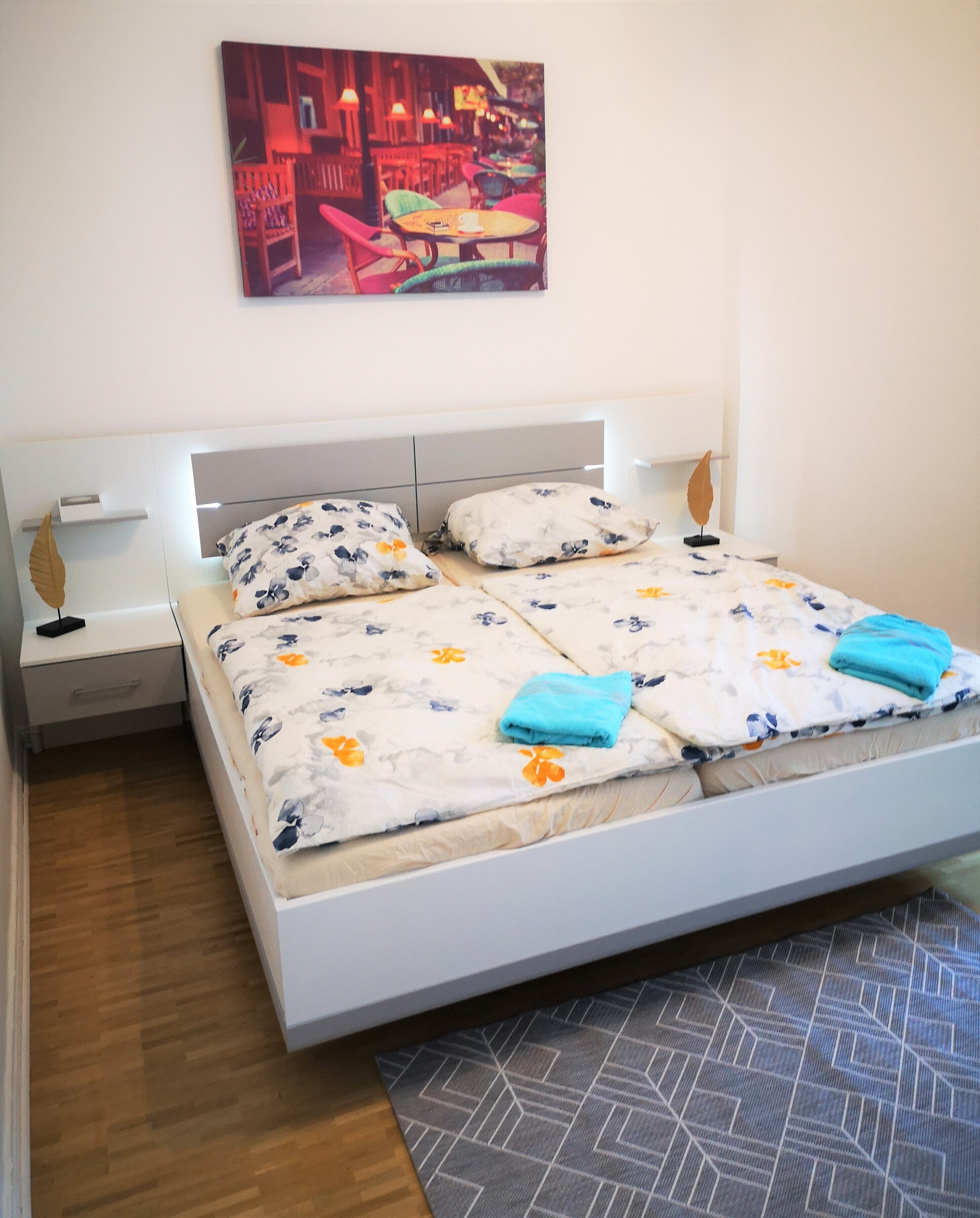 A neatly made bed with floral bedding and two blue towels, a bedside table with a lamp, and a painting of a café scene above the bed.