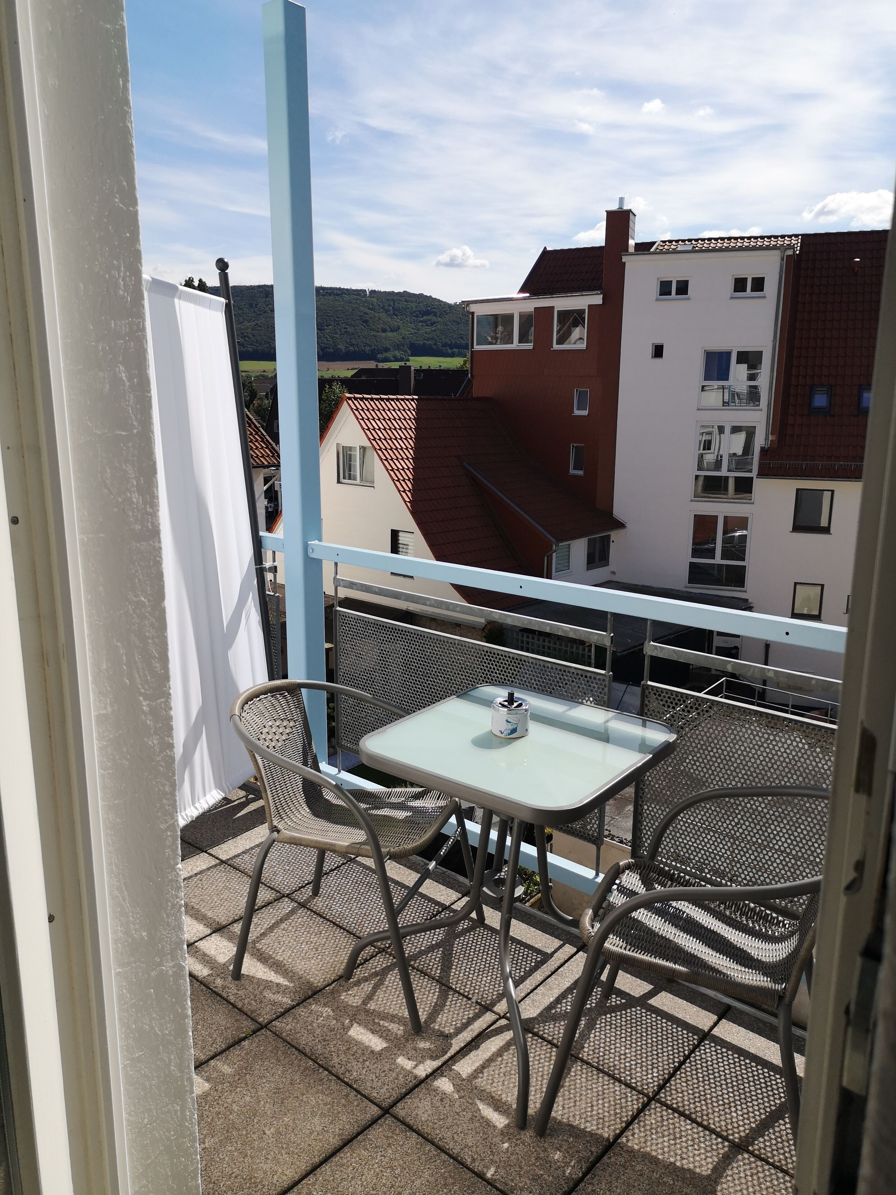 Un balcon avec une table et des chaises, donnant sur un quartier résidentiel avec des maisons et un paysage vallonné.