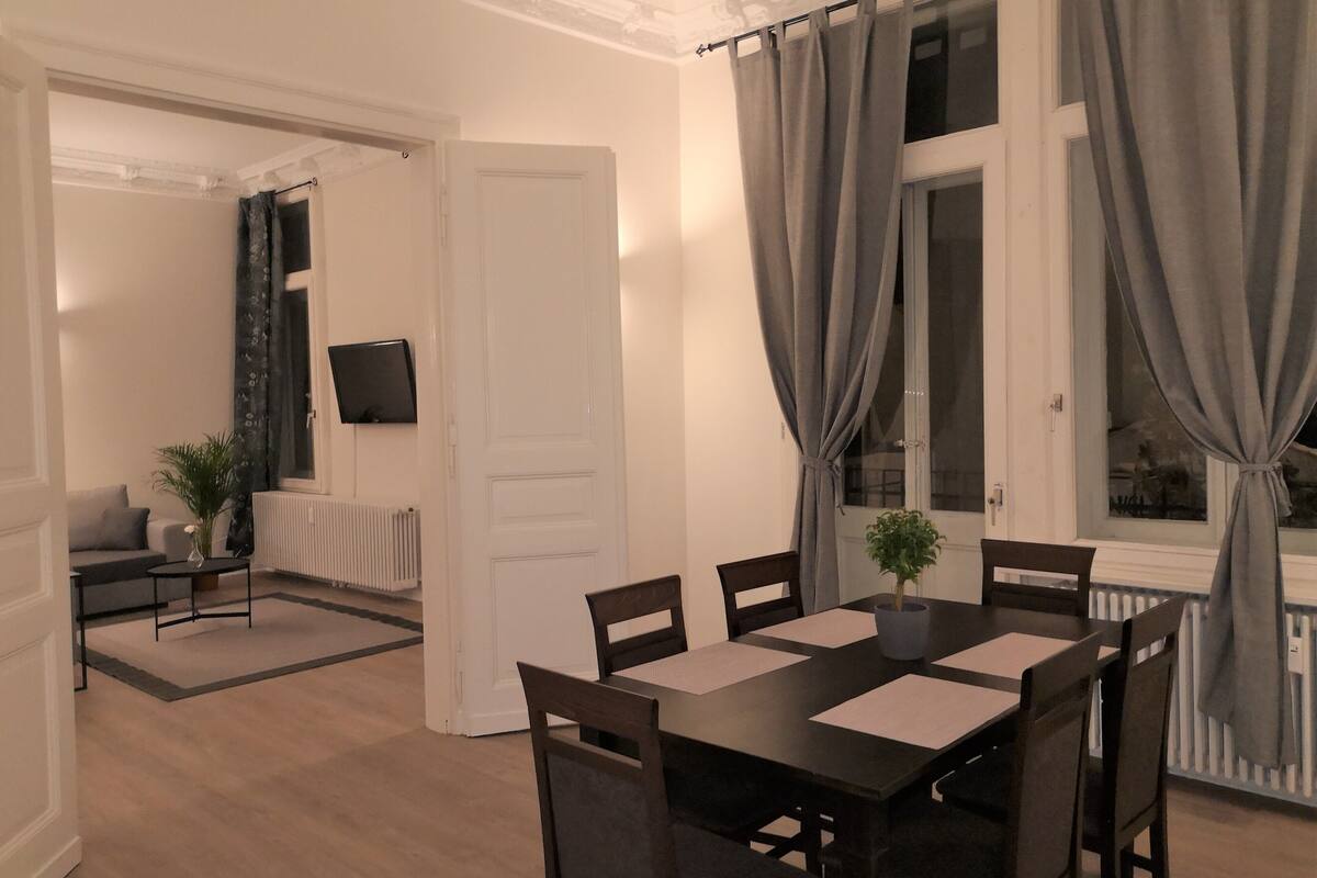 A dining area with a table and chairs, a television on the wall, and a view of the outdoors through a window with curtains.