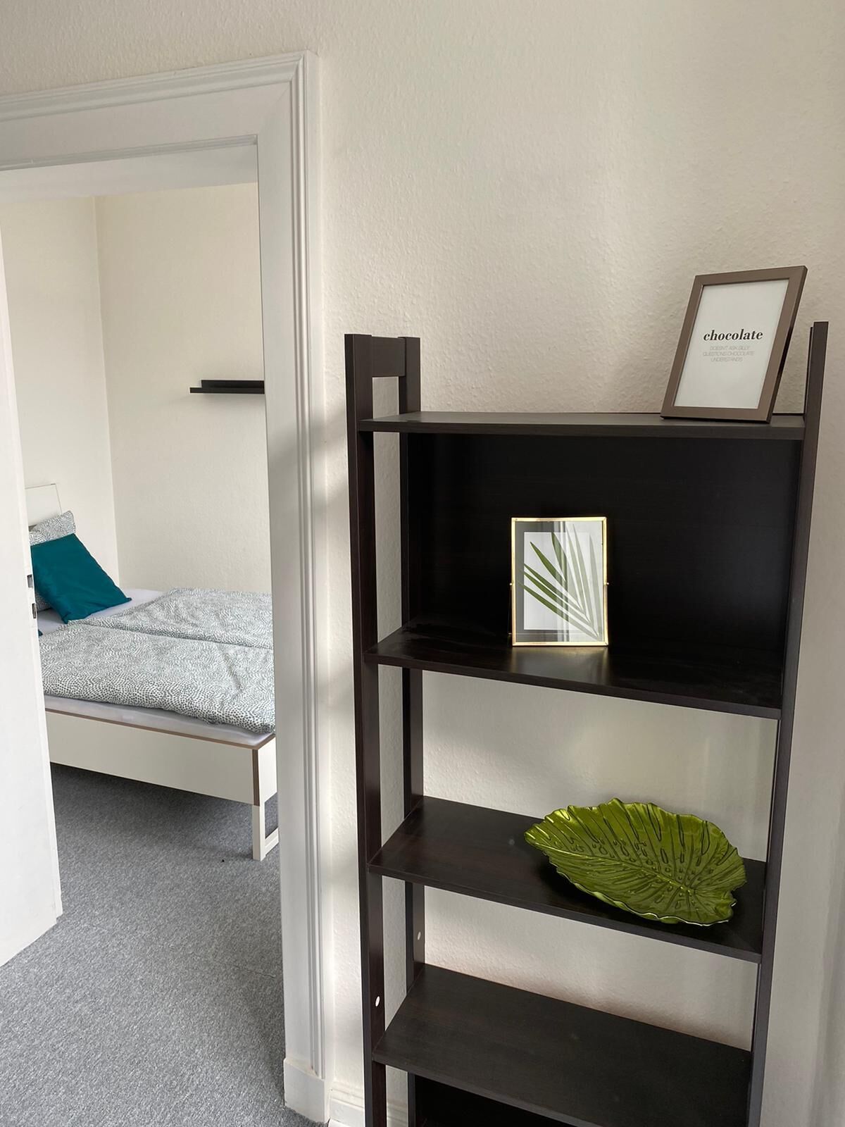 A bedroom with a bed, a dark wooden shelf unit with a framed picture and a green decorative item, and a white door leading to another room.