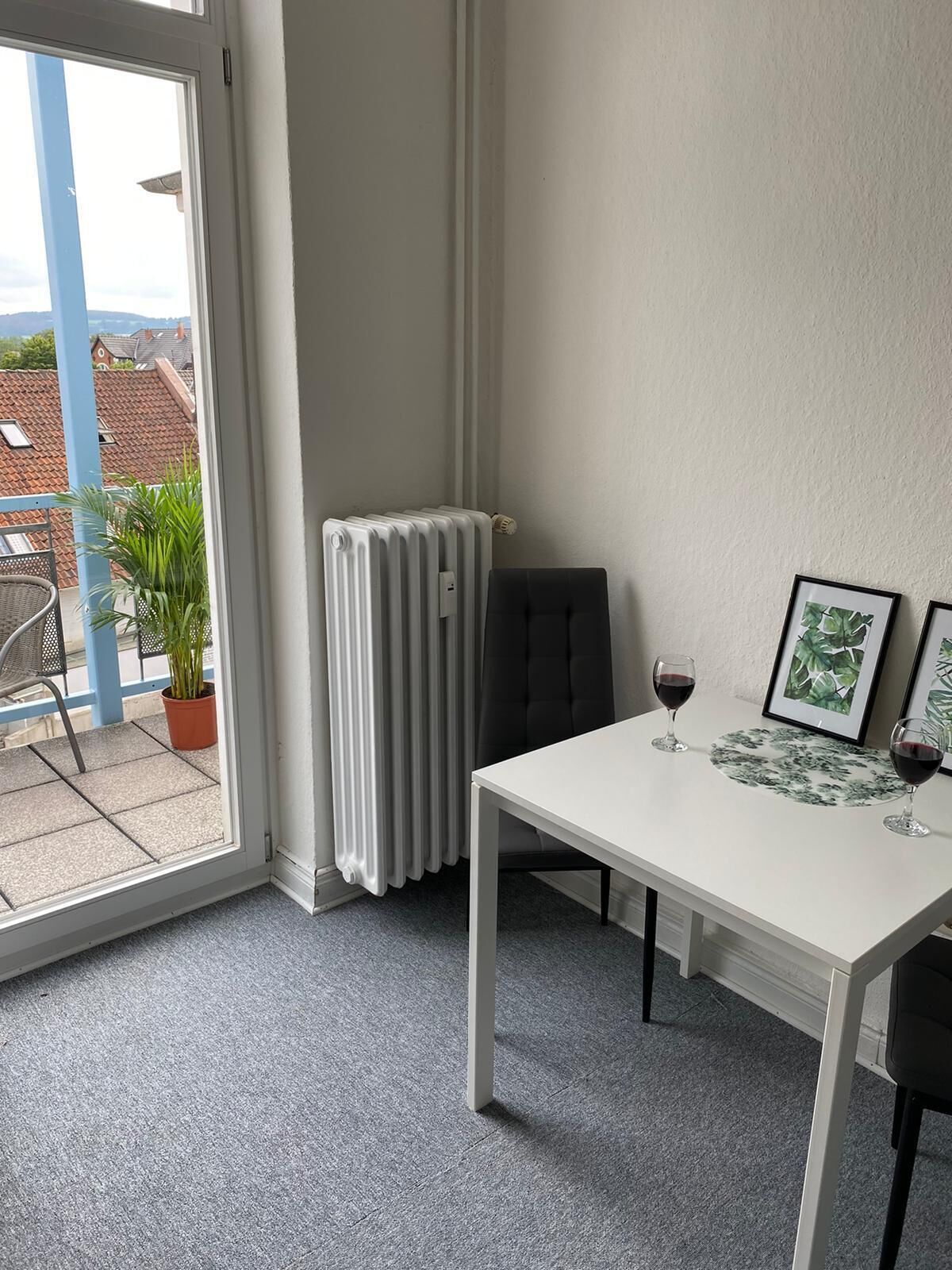 Una habitación con una mesa blanca, dos sillas, un radiador, una planta en maceta y cuadros en la pared.