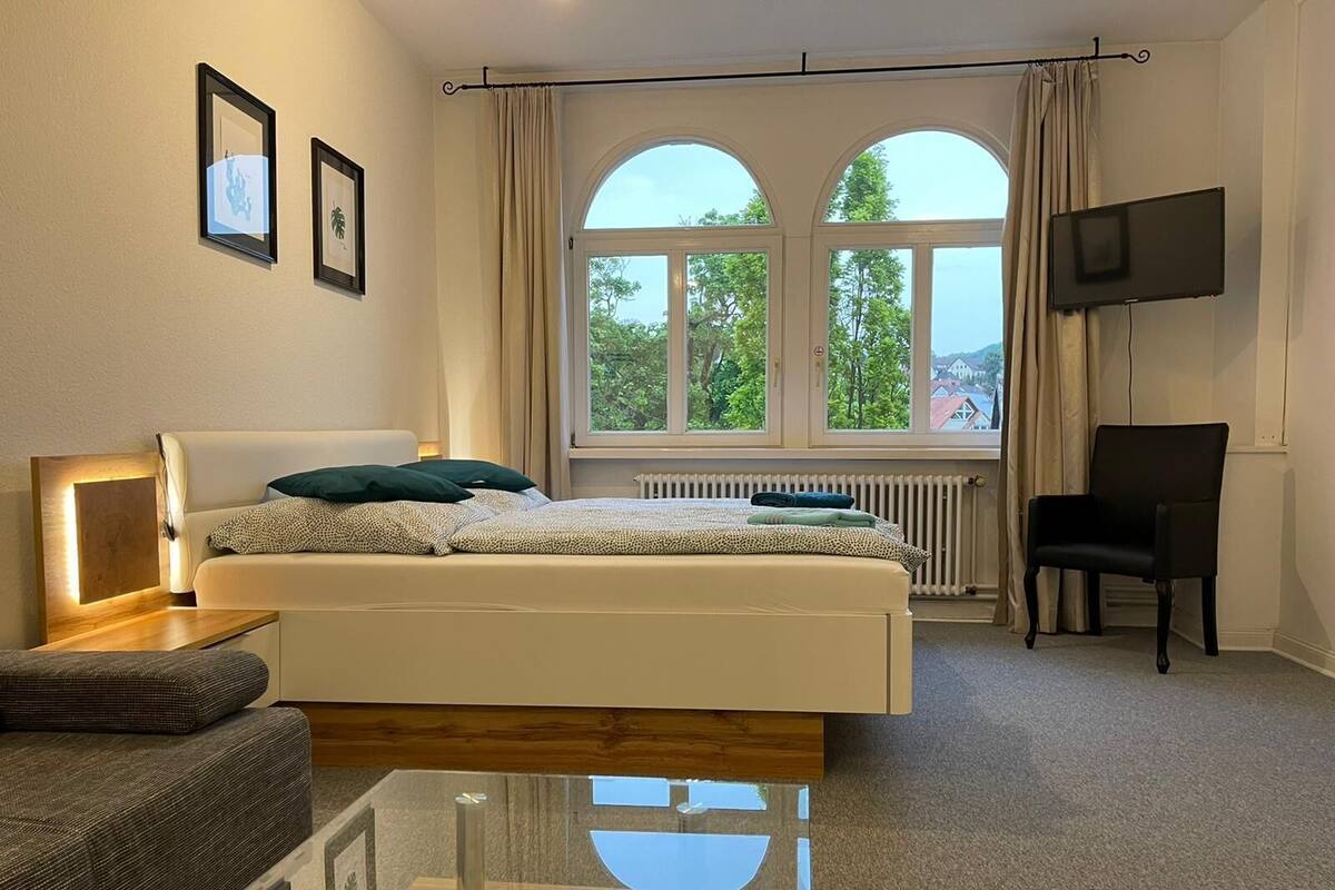 A bedroom with a bed, a chair, a TV, and a window with a view of trees.