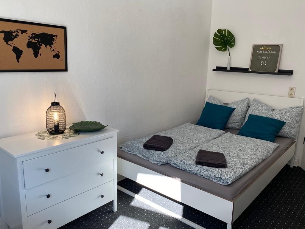 A neatly arranged bedroom with a bed, a bedside table with a lamp, a world map on the wall, and a welcome sign.