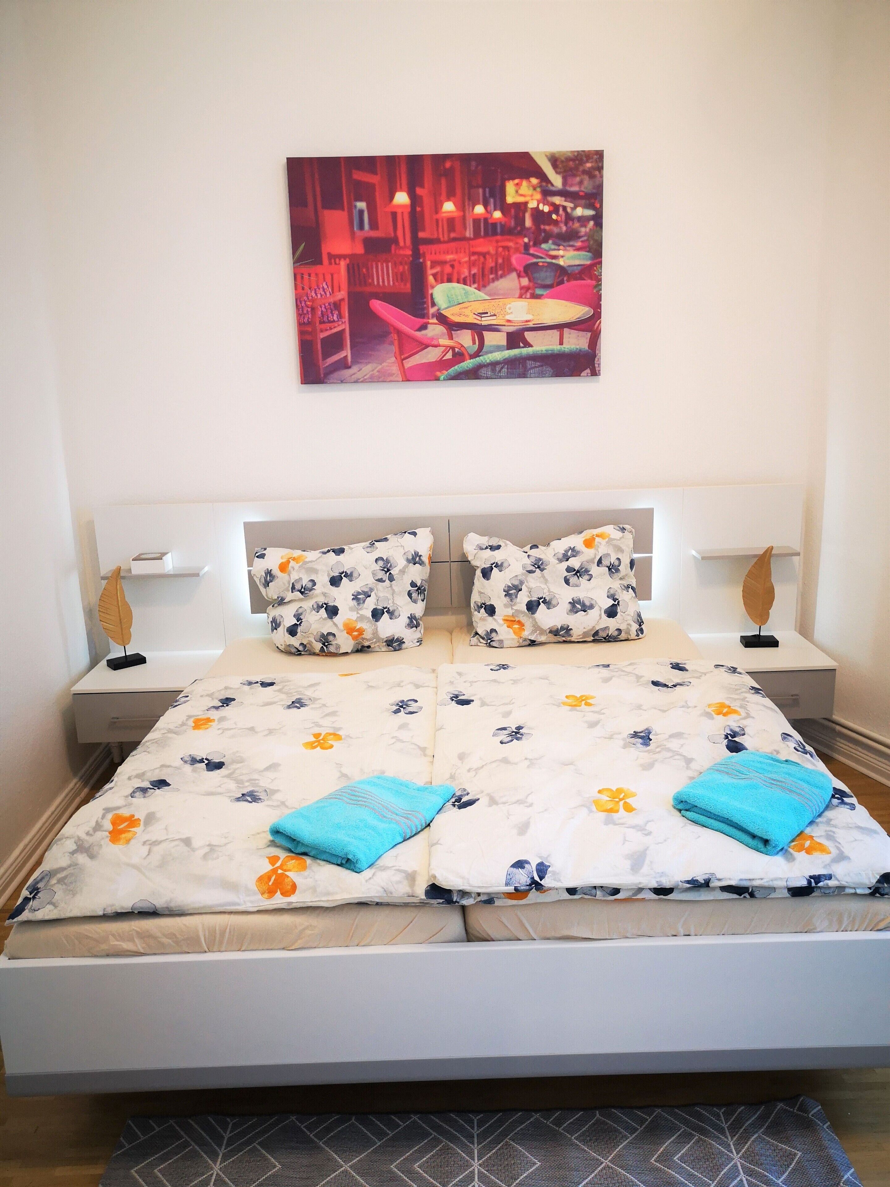Una cama bien hecha con ropa de cama floral, dos almohadas y dos toallas dobladas. Hay dos mesitas de noche con lámparas, una obra de arte en la pared y un pequeño objeto decorativo en la mesita de noche.