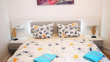 Una cama bien hecha con ropa de cama floral, dos almohadas y dos toallas dobladas. Hay dos mesitas de noche con lámparas, una obra de arte en la pared y un pequeño objeto decorativo en la mesita de noche.