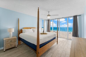 Un letto a baldacchino con struttura in legno, un comodino con cassetti, una lampada e vista sulla spiaggia attraverso ampie finestre.