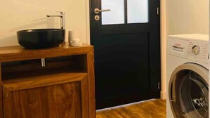 Un baño pequeño con un tocador de madera, un espejo redondo, una puerta negra y una lavadora de carga frontal.