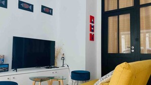 Una sala de estar con un televisor de pantalla plana grande, un sofá amarillo, un reposapiés azul y una pequeña mesa de vidrio.