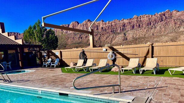 A pool area with clear blue water, lounge chairs, and a wooden fence with a 'No Swimming' sign. - T4 Corner Moab Retreat with Private Garage in Rim Village (Moab)