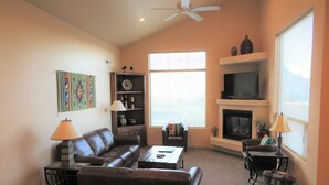 A living room with a fireplace, a ceiling fan, and a large window. - T4 Corner Moab Retreat with Private Garage in Rim Village (Moab)