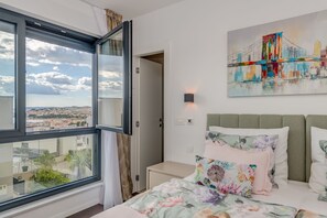 A bedroom with a large window, a bed with floral bedding, a cityscape painting, and a nightstand.