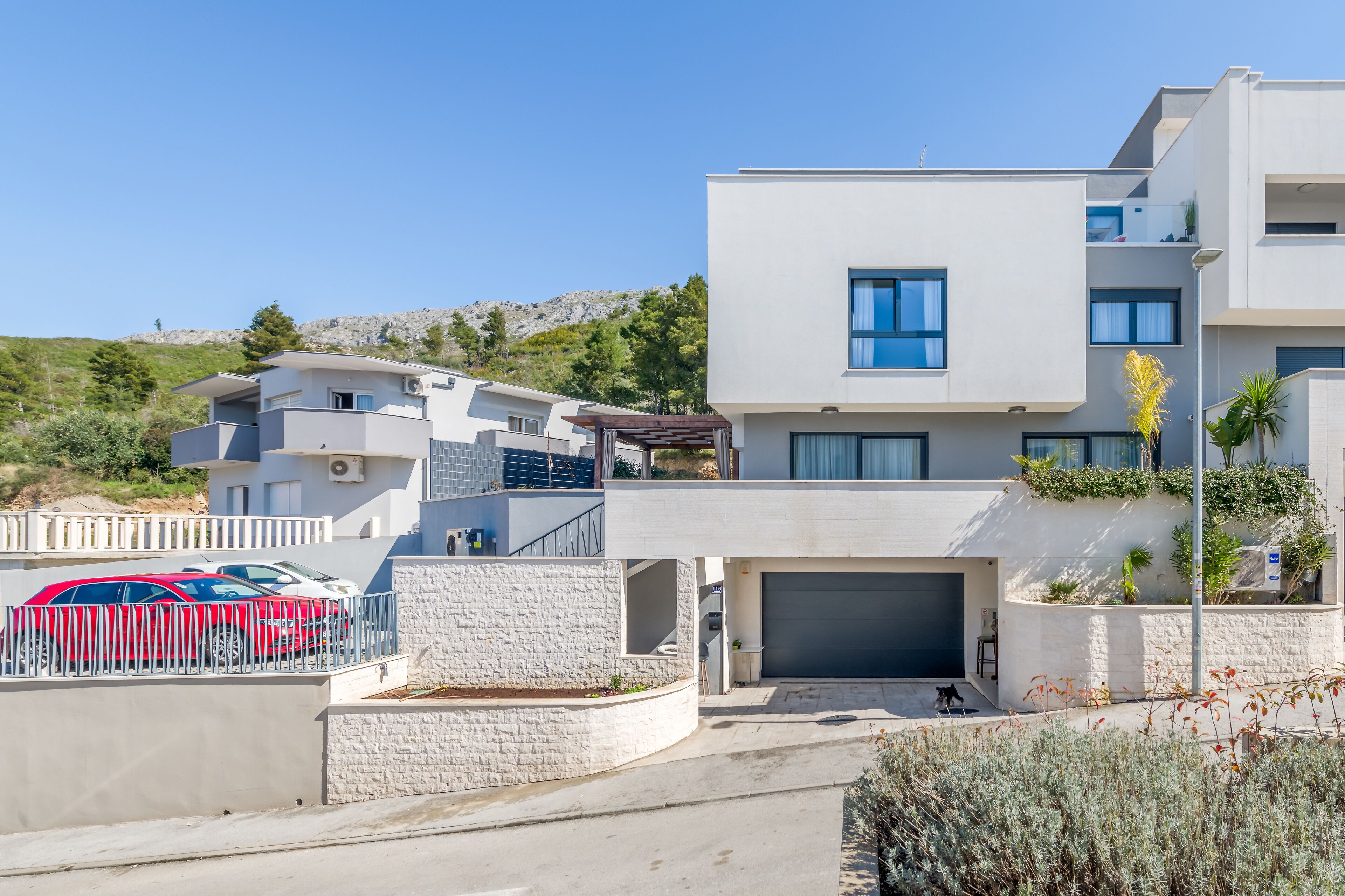 Un bâtiment moderne à deux étages, doté d’un garage, avec une voiture rouge garée devant et une colline en arrière-plan.