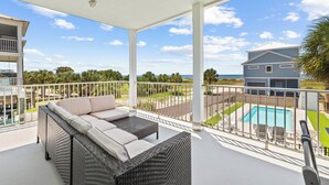 A covered balcony with wicker furniture, a pool, and a view of the ocean and neighboring buildings. - One Wave or Another: Gulf Views, Private Pool, Putting Green (Indian Pass)