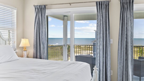 A bedroom with a large bed, a nightstand, a lamp, and a view of the ocean through a sliding glass door. - One Wave or Another: Gulf Views, Private Pool, Putting Green (Indian Pass)