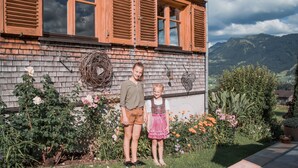 A house with wooden shutters, a garden with roses, and a mountainous backdrop.