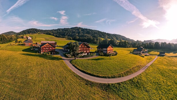 A rural landscape with a winding road, several traditional houses, and a backdrop of rolling hills and a clear sky.