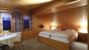 A wooden cabin room with a bed, a table with chairs, and a window with curtains.