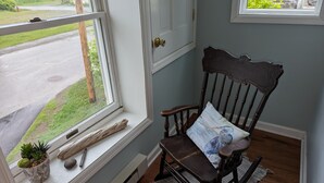 Une pièce avec un fauteuil à bascule en bois, une fenêtre donnant sur une route et sur la verdure, une petite plante posée sur le rebord de la fenêtre, et une porte avec une poignée en laiton.