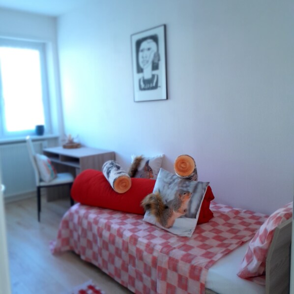 Une petite chambre à coucher, bien rangée, avec une couvre-lit à carreaux, un oreiller rouge et un tableau accroché au mur.