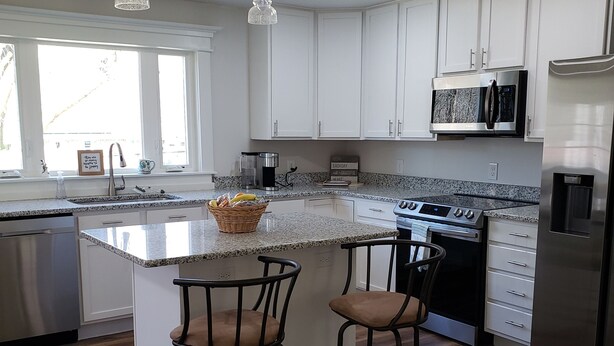 A modern kitchen with white cabinets, a granite countertop, a stainless steel refrigerator, a microwave, and a gas stove. There are two bar stools and a window providing natural light. - Creekside cottage home near Red River Gorge and Natural Bridge State Park (Stanton)