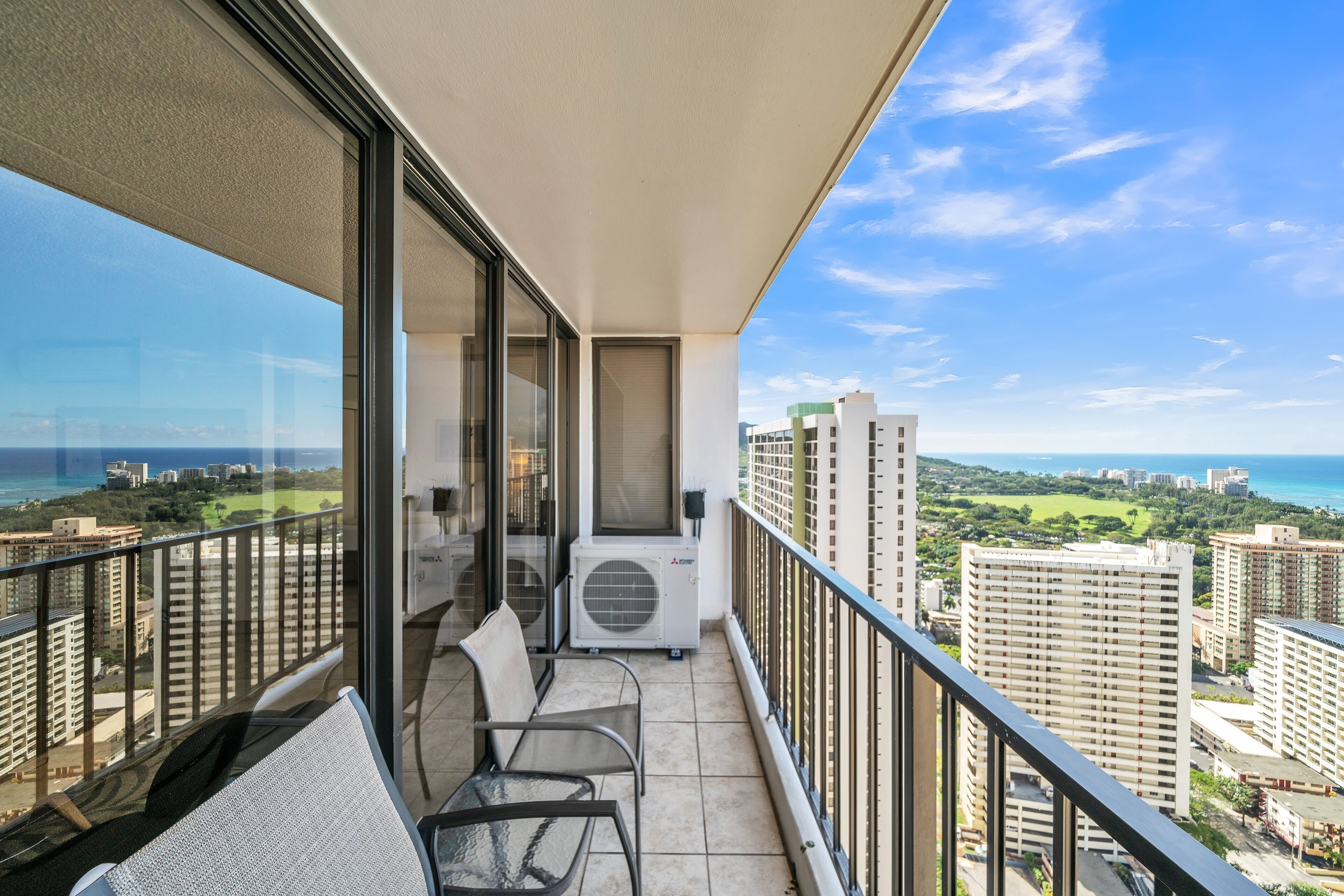 A balcony with a view of the city and ocean, featuring a seating area and an air conditioning unit.