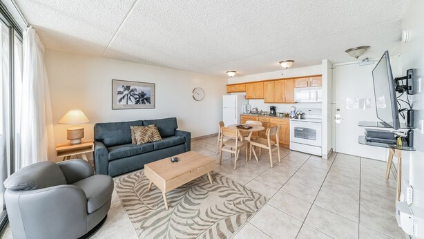 A modern living room with a sofa, armchair, coffee table, and a dining area with a table and chairs. There is a kitchen with cabinets and appliances, and a TV mounted on the wall. - Waikiki Banyan Condo With Panoramic 37th Floor Ocean Views (Honolulu)