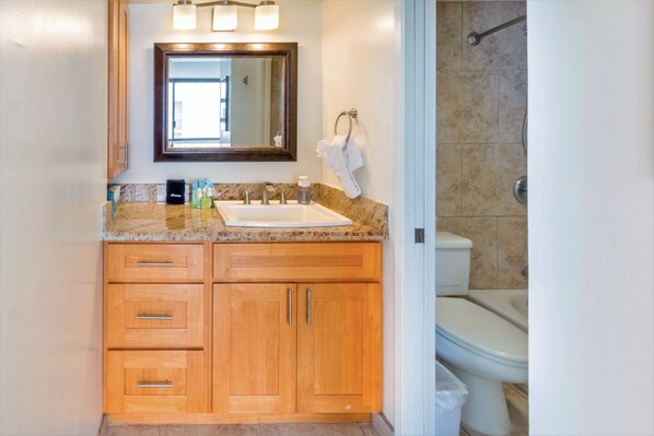 A bathroom with a marble countertop, wooden cabinets, a sink, a toilet, and a shower area. - Waikiki Banyan Condo With Panoramic 37th Floor Ocean Views (Honolulu)