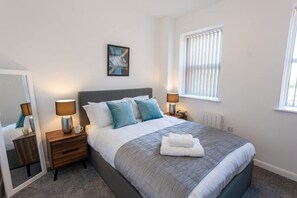 A bedroom with a bed, bedside tables, lamps, a window with blinds, a painting on the wall, and a radiator. - Modern 2 Bedroom Bolton Apartment with WiFi (Bolton)