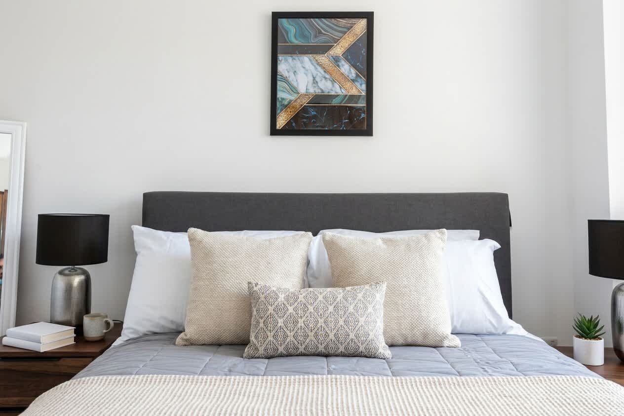 A neatly made bed with a grey headboard, white and patterned pillows, and a framed abstract painting above.
