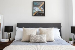 A neatly made bed with a grey headboard, white and patterned pillows, and a framed abstract painting above. - Modern 2 Bedroom Bolton Apartment with WiFi (Bolton)