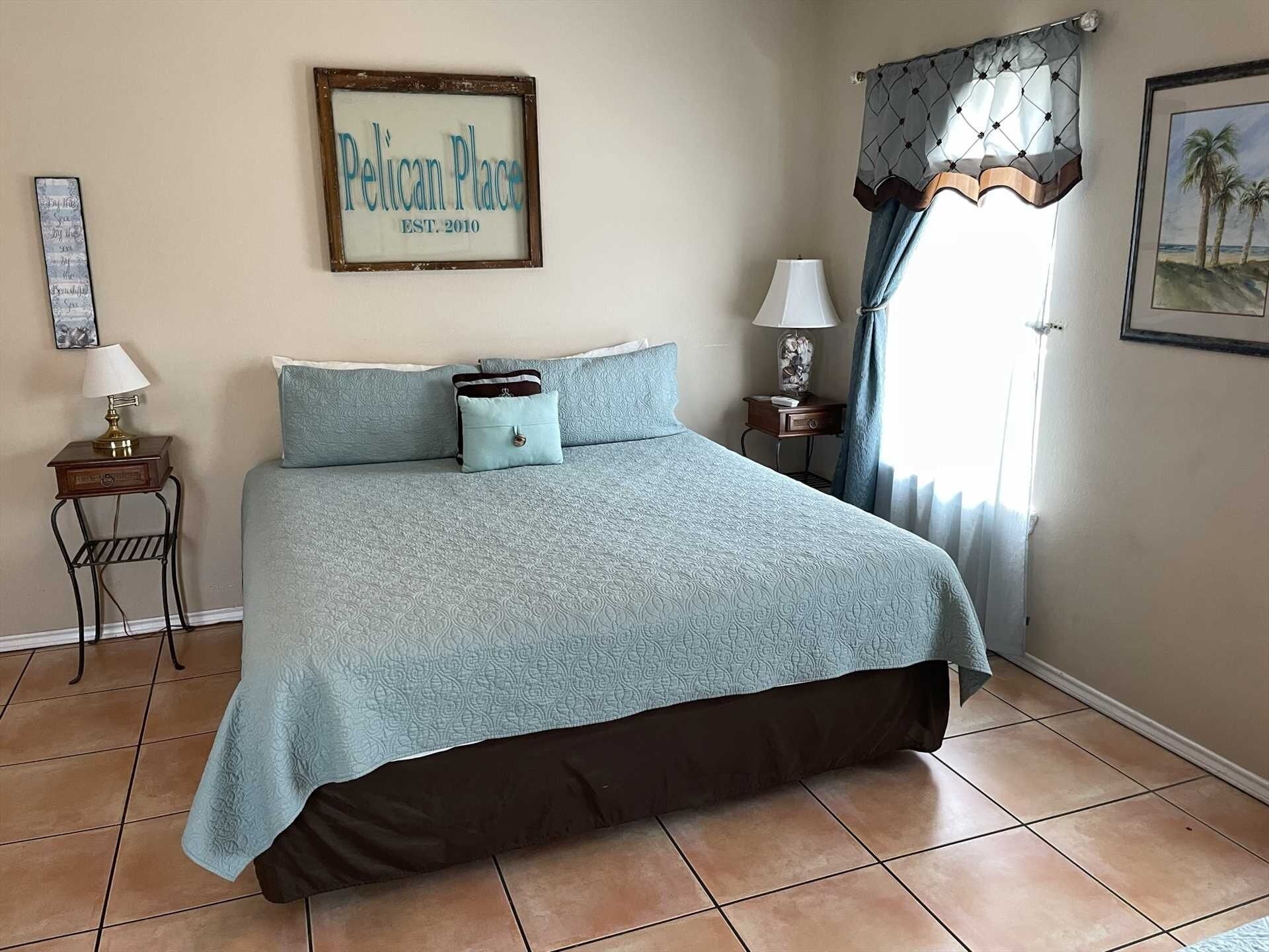 A bedroom with a bed, two bedside tables, a lamp, a framed sign, and a window with curtains.