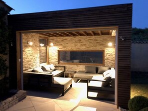An outdoor patio with a stone wall, wooden beams, and a large window. There are several seating areas with cushions and a table.
