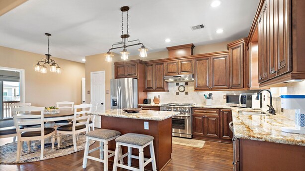 A spacious kitchen with wooden cabinets, a central island, stainless steel appliances, and a dining area with white chairs and a table. - Copper Top | Moments from Beach (Galveston)