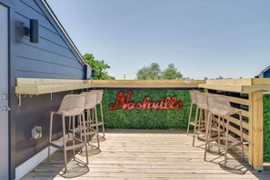 Un área de estar al aire libre con dos sillas de mimbre y una mesa de madera, una pared verde con la palabra 'Nashville' escrita y un cielo despejado.