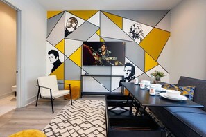 A modern living room with a flat-screen TV mounted on a geometric-patterned wall, a sofa, a dining table with a few items on it, and a chair. - Nashville getaway: themed house with garage game room, sleeps 12 (Nashville)
