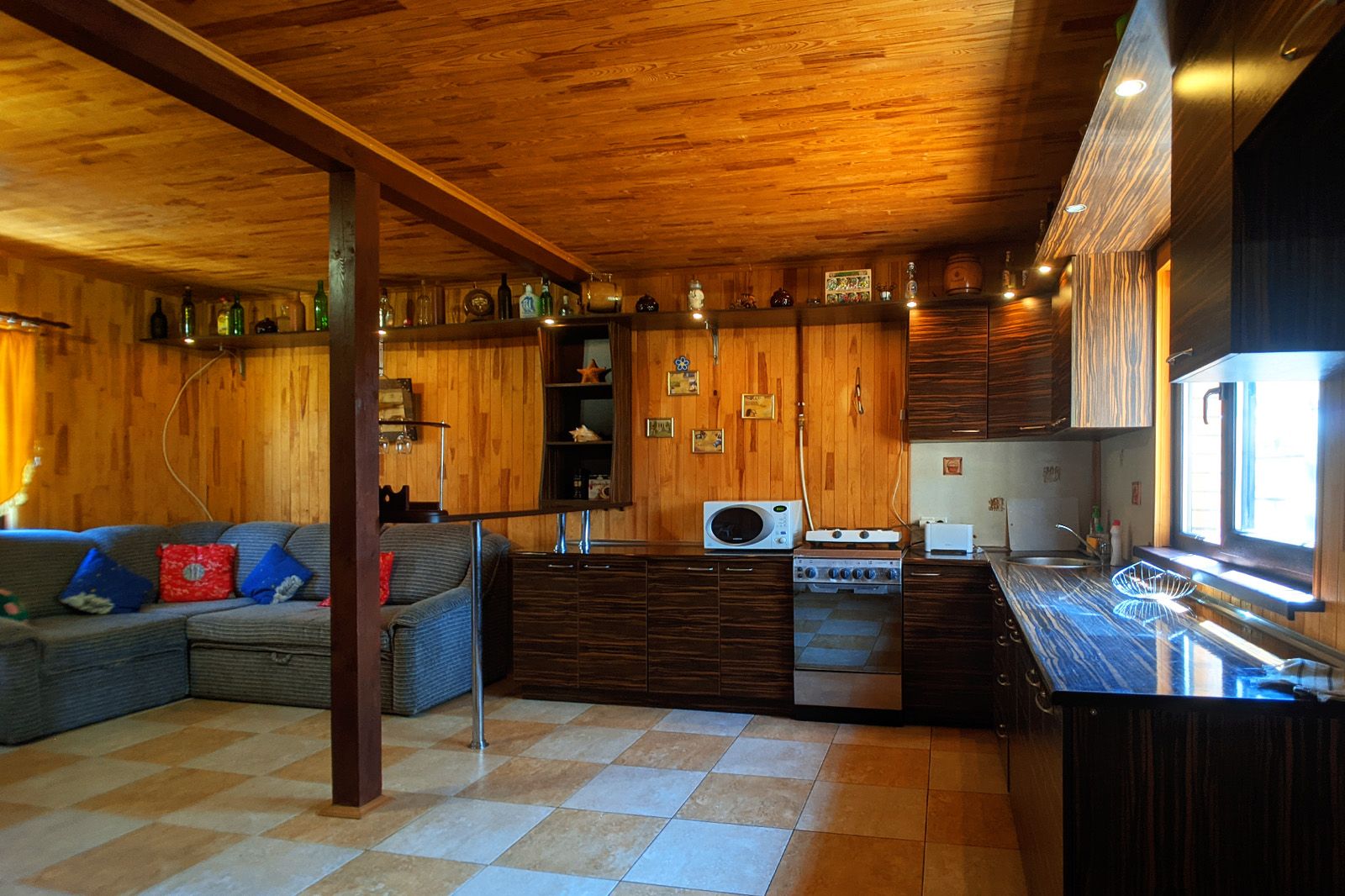Cozinha compacta com armários de madeira, micro-ondas, fogão e pia. Na área de estar, há um sofá com almofadas.