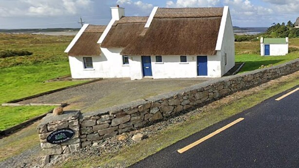 Ein traditionelles weißes Cottage mit einem Strohdach, einer Steinmauer davor und einem Schild, das anzeigt, dass es Teil eines Dorfes ist.