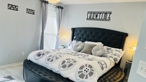 A modern bedroom with a black upholstered bed, white bedding with film reel patterns, a round black ottoman, and a wall-mounted light fixture. - 4 Miles from Disney Hollywood Themed Home (Kissimmee)