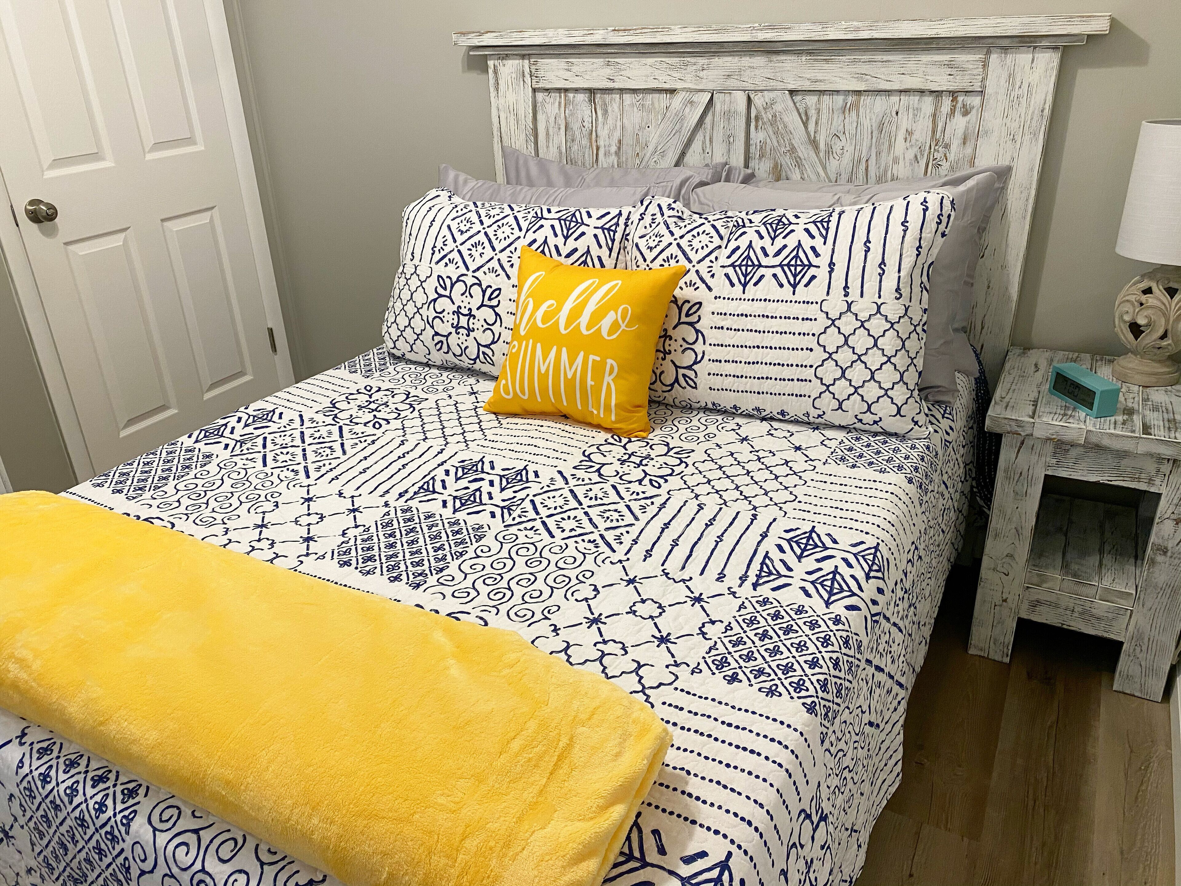 Un lit avec une literie à motifs, un plaid jaune et un coussin orné de l’inscription « bonjour, été ».