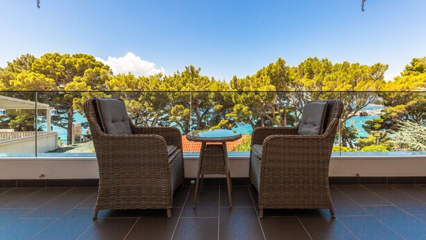 Varanda com duas cadeiras de vime e uma pequena mesa redonda, com vista para uma piscina e árvores.
