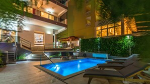 Uma piscina com espreguiçadeiras, escada e um edifício com varandas.